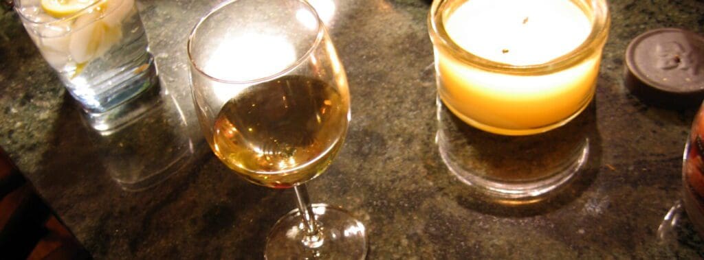 A glass of white wine and a lit candle on a dark table.