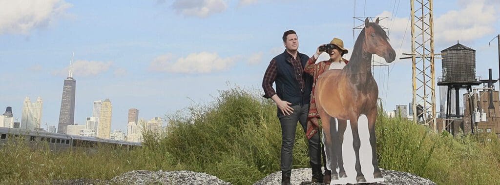Two people outdoors, one in a dark shirt and the other in a hat, walking near bushes.