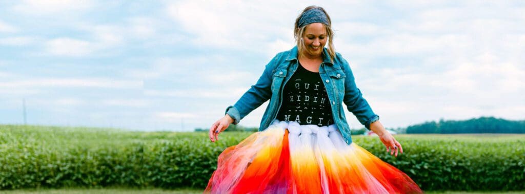 A smiling woman wearing a colorful rainbow skirt and denim jacket outdoors.