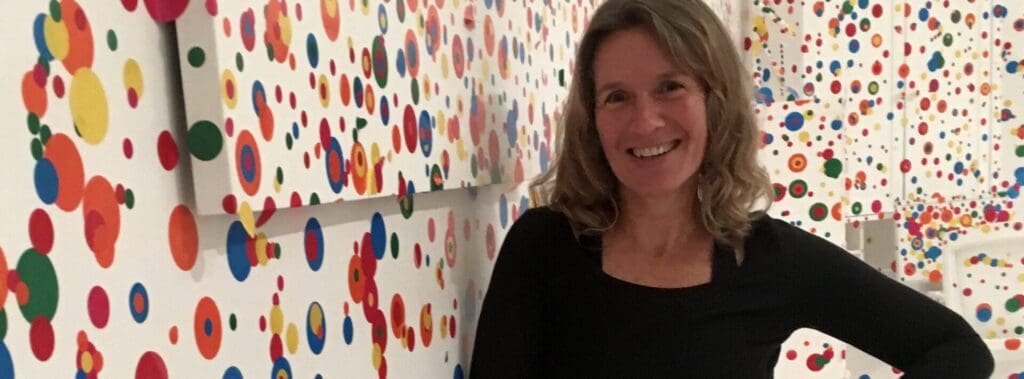Smiling woman standing next to colorful abstract art with vibrant dots.
